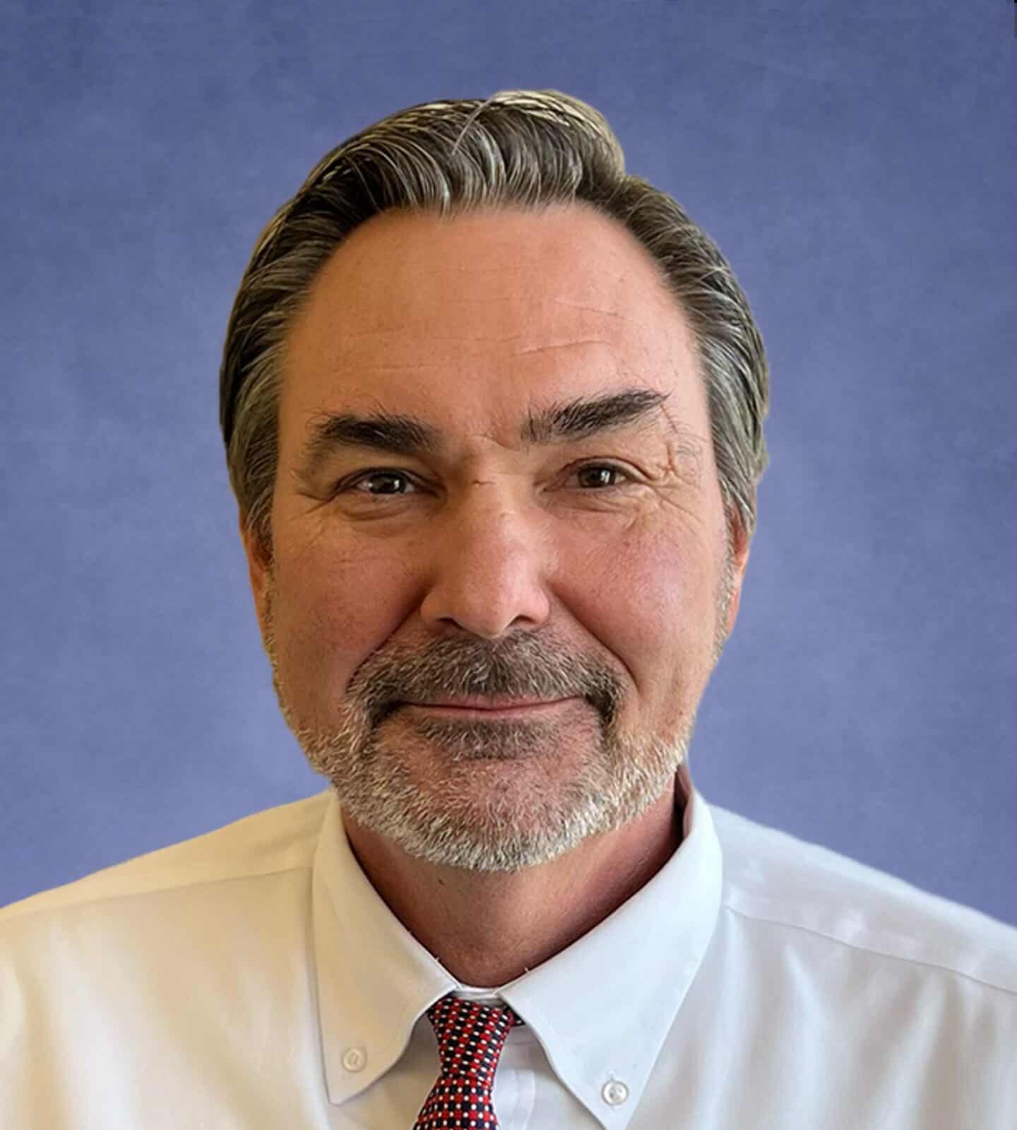 Professional portrait of a middle-aged man with gray hair and beard, wearing a white shirt and red-tie.