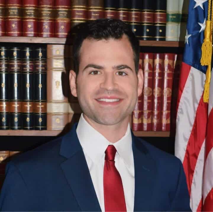 DCFS Public Affairs Director Lester Duhé in a blue suit with a red tie, standing in front of a bookcase with the American flag to his left.